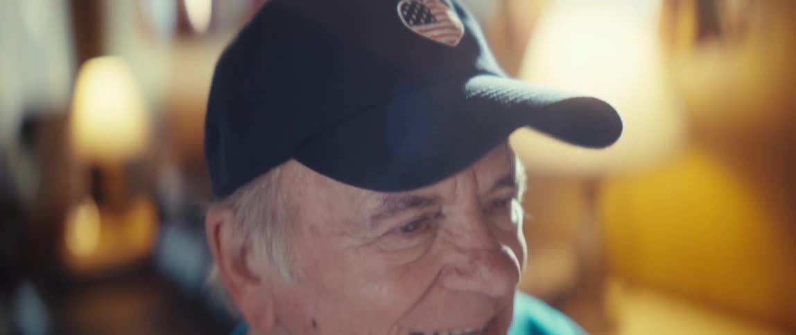 Elderly man wearing a baseball cap and a soccer shirt smiling and gesticulating indoors