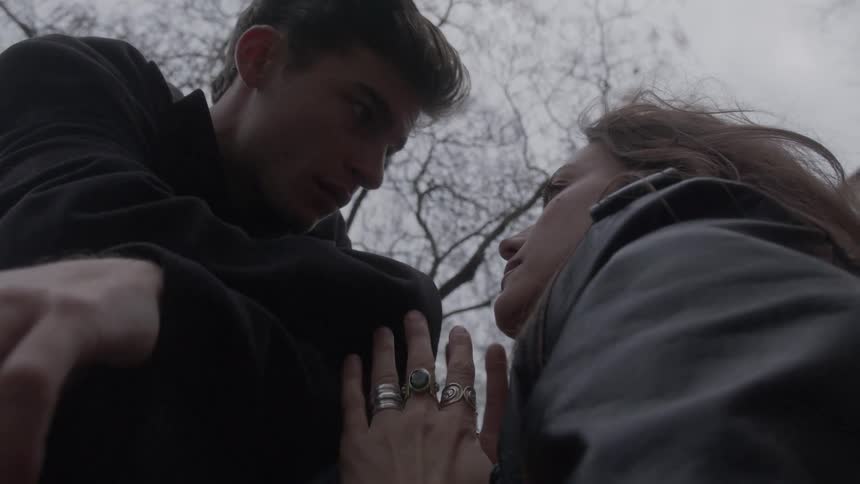 A young couple sits in the front yard of a building hugging and kissing while wearing jackets during the fall season with the trees bare of leaves