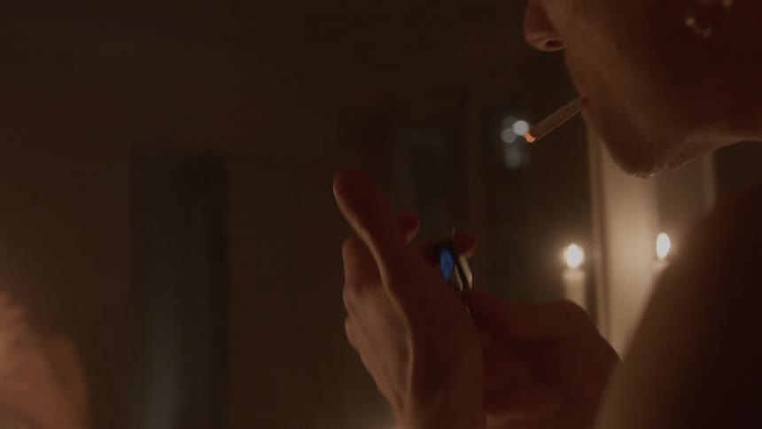 A man lights a cigarette then looks in the mirror in a candle lit room