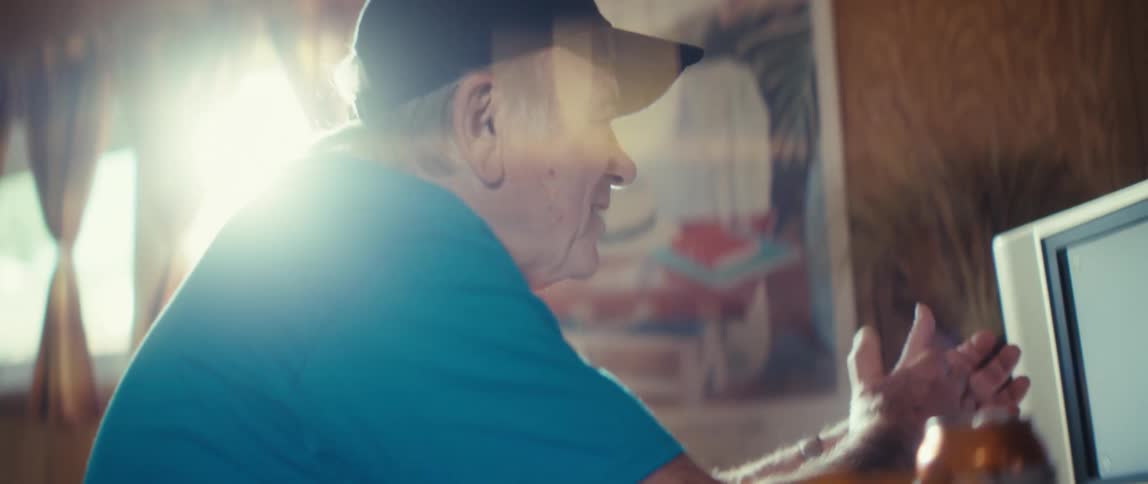 Elderly man in a blue shirt and dark cap claps while looking at a screen in a woodpaneled room with natural light