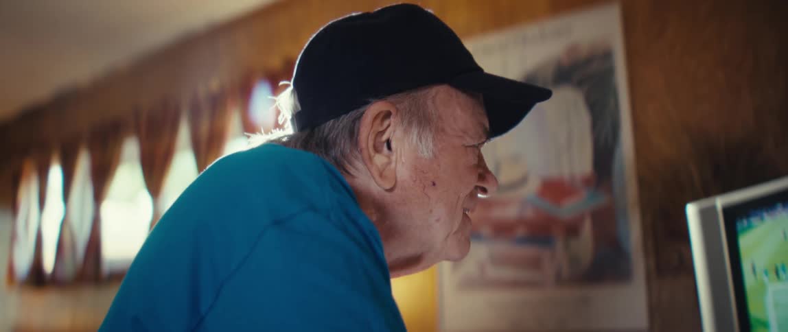 A closeup profile shot captures an elderly man in a blue tshirt and a black baseball cap looking down and smiling