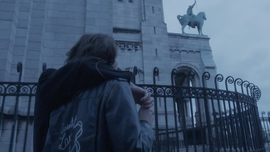 Couple embraces near an iron fence with a historic building and statue in the background under a cloudy sky