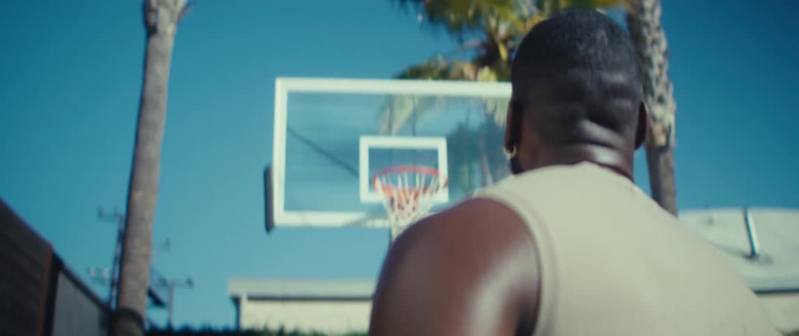 An overtheshoulder shot focused on a man wearing a tan tank top and a hoop earring prepares to engage with the game while he focuses on a clear rectangular glass basketball backboard with an orange rim and white net set against a vibrant blue sky