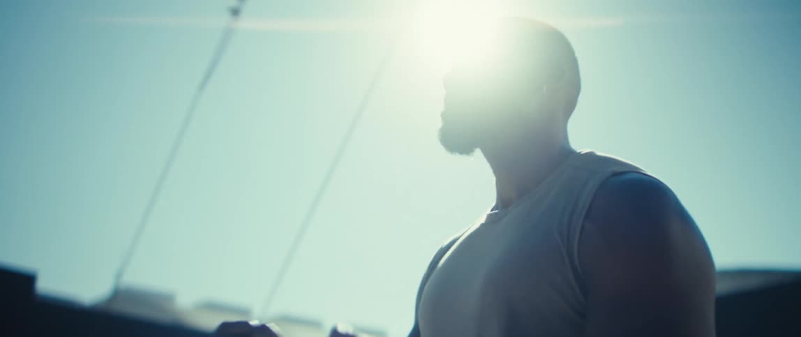 A focused man with a full beard as he runs or jogs forward on a bright clear day in an urban setting the bright sun flaring in the frame while the background reveals distant buildings and utility poles
