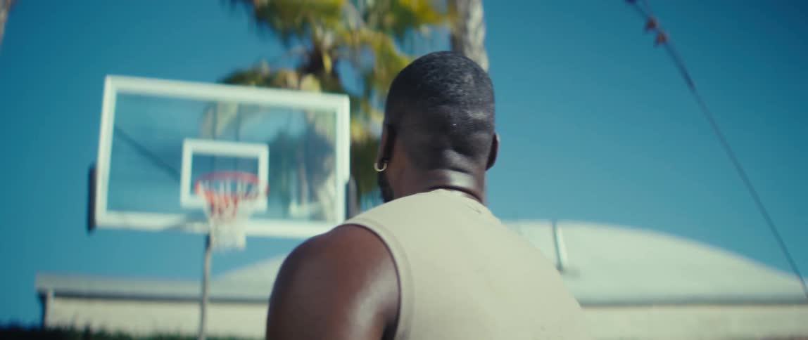 A man in a sleeveless shirt faces a backyard basketball hoop under a clear blue sky with palm trees