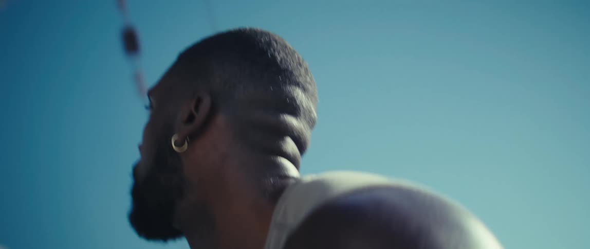 A man in a sleeveless shirt begins by looking offcamera on a bright sunny day with the light creating a lens flare and silhouetting his figure against the sky he then quickly turns his head and body to his right revealing a clear blue sky a palm tre