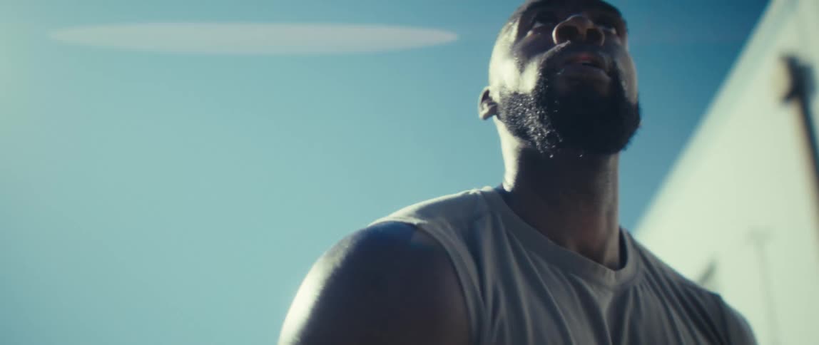 A muscular man in a sleeveless shirt walks and dribbles a basketball under bright sun against a clear blue sky