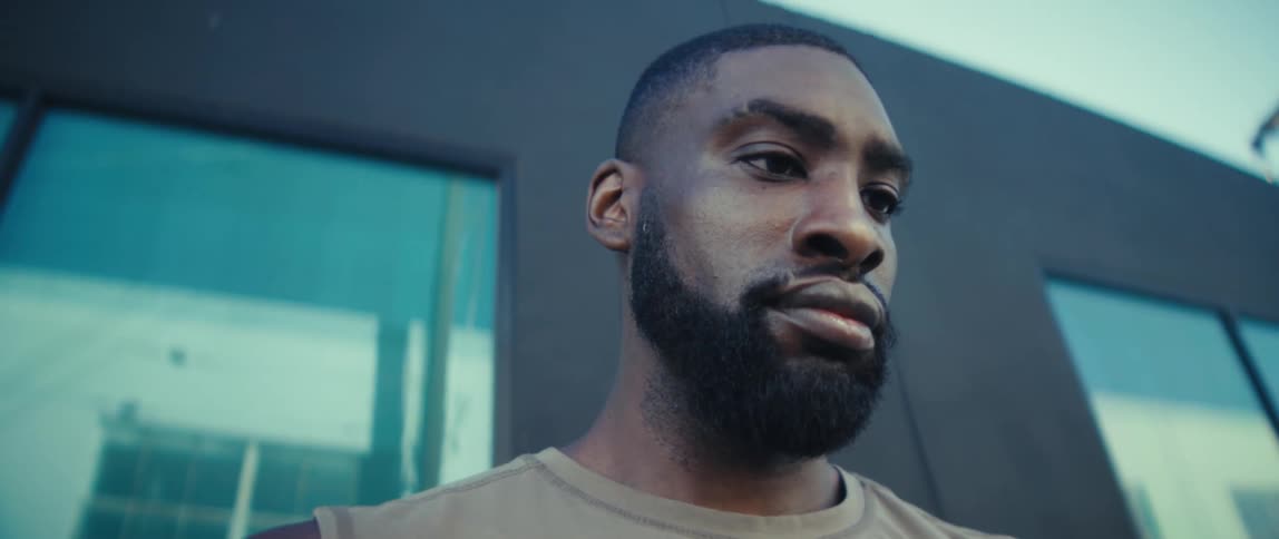 A young black man with a beard stands outdoors at midday looking at his phone