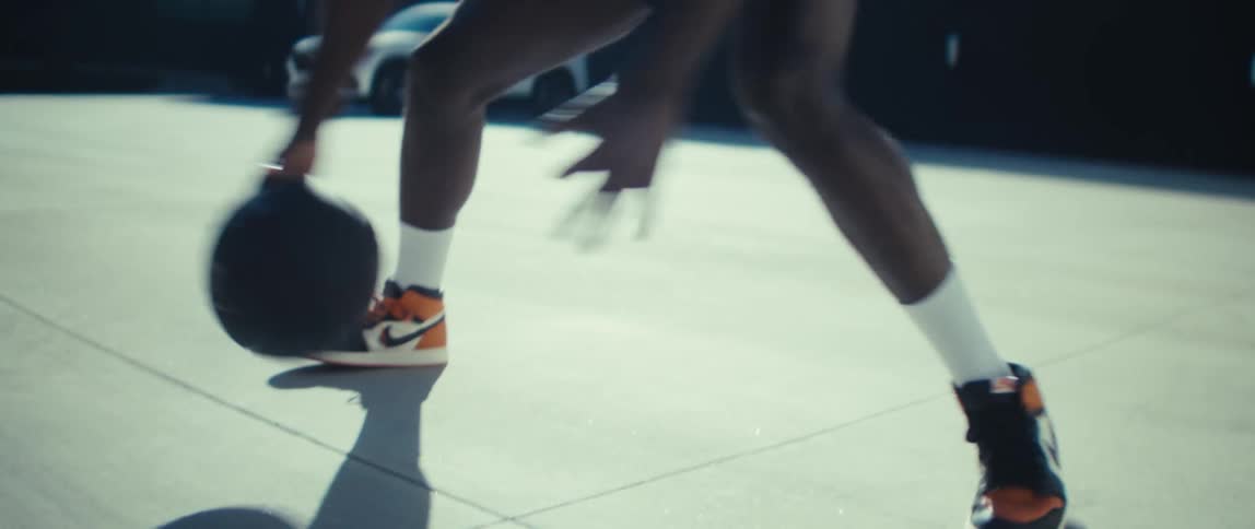 A black man in white socks and orange and black sneakers dribbles a basketball low on a sunlit outdoor court