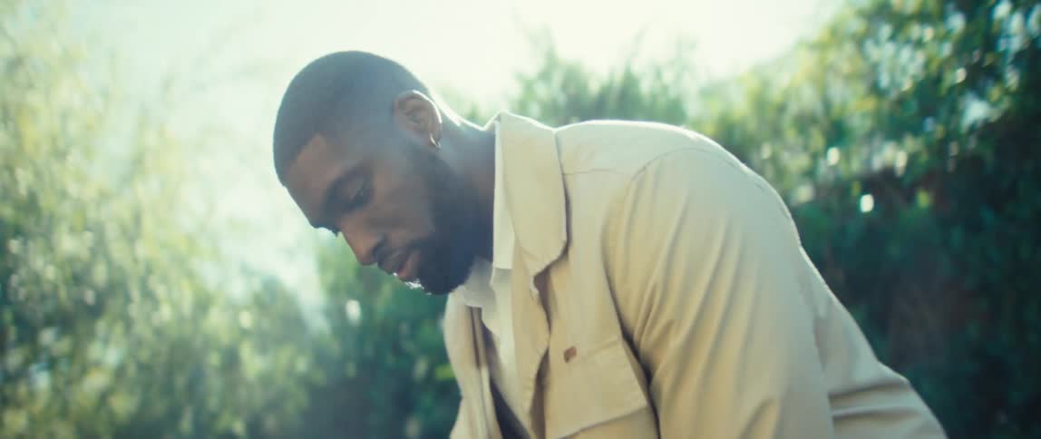 A man in a light beige jacket leans forward in soft sun near a hedge looking down with a focused expression
