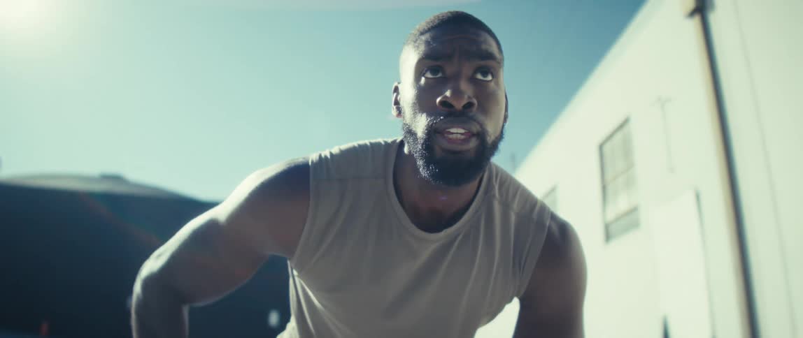 A focused black man in a sleeveless shirt crouches in bright sunlight as he readies to play with a ball with city buildings blurred behind him