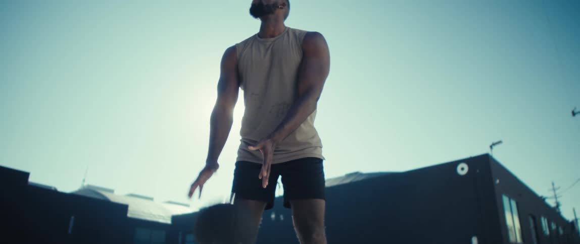 A muscular man in a sleeveless shirt dribbles and shoots a basketball under bright sun looking upward with a focused expression against a clear blue sky