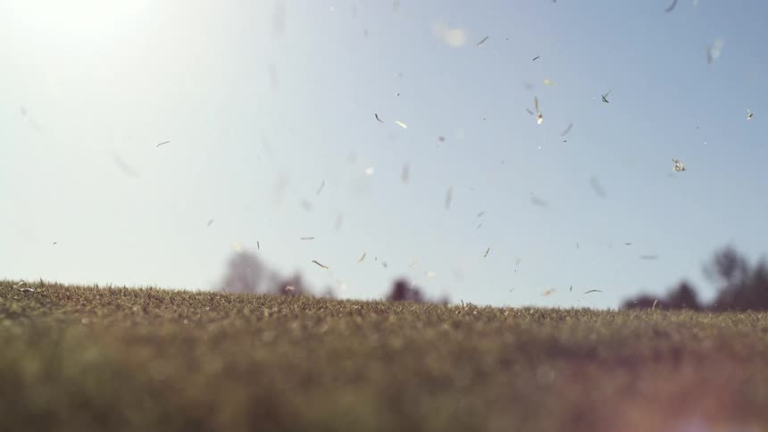 A golf club swings down sharply and strikes a golf ball near the ground simultaneously kicking up a large dramatic spray of dirt and grass that fills the frame the debris hangs in the air against a bright hazy sky slowly drifting down while the ball