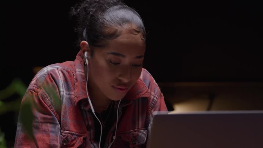 A young woman in a darkened room with an opened laptop in front of her and wearing earphones nods intently as she uses a pen to write something