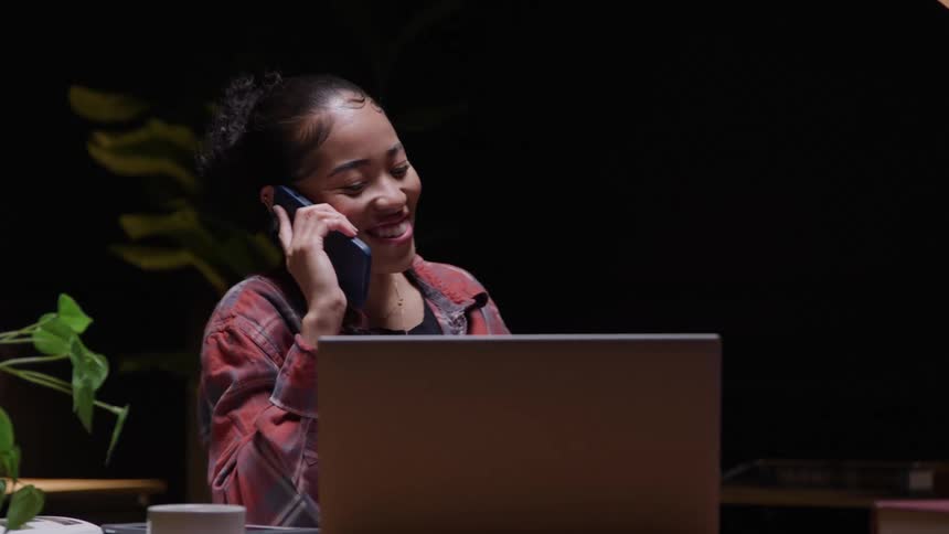 A young woman laughs while sitting at a work desk and talking on her smartphone