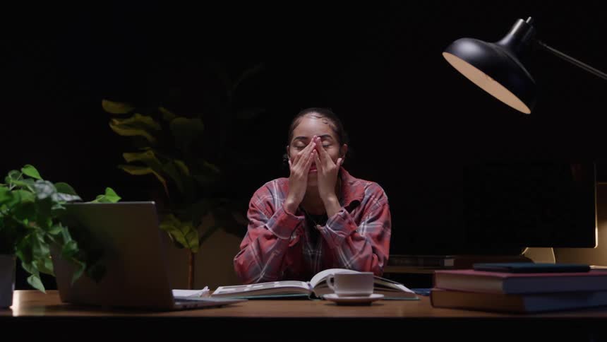 Young woman sitting at a desk with a laptop computer and studying a book pauses to rub her face and rest her head in her hands