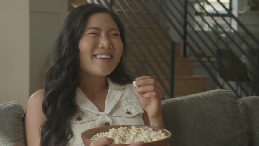 Young asian woman sitting down on a couch laughing and eating popcorn from a bowl indoors during day time