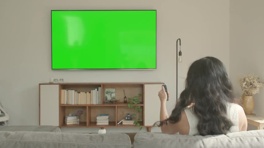 Woman carrying a bowl of popcorn sits on her couch and turns on the television