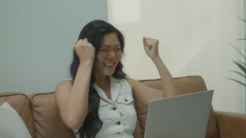 Woman sitting on a couch cheers and claps after getting good news via her laptop