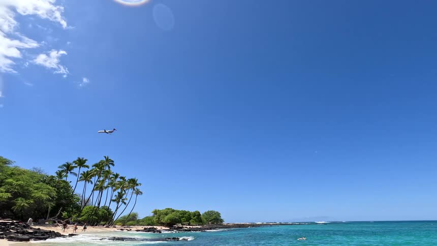 A tropical beach with tall palm trees and turquoise water under a clear blue sky as a lowflying airplane passes overhead while a few people relax on the sand and swim