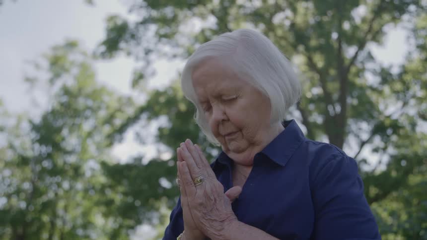 An elderly woman bows her head while praying outdoors on a summer day