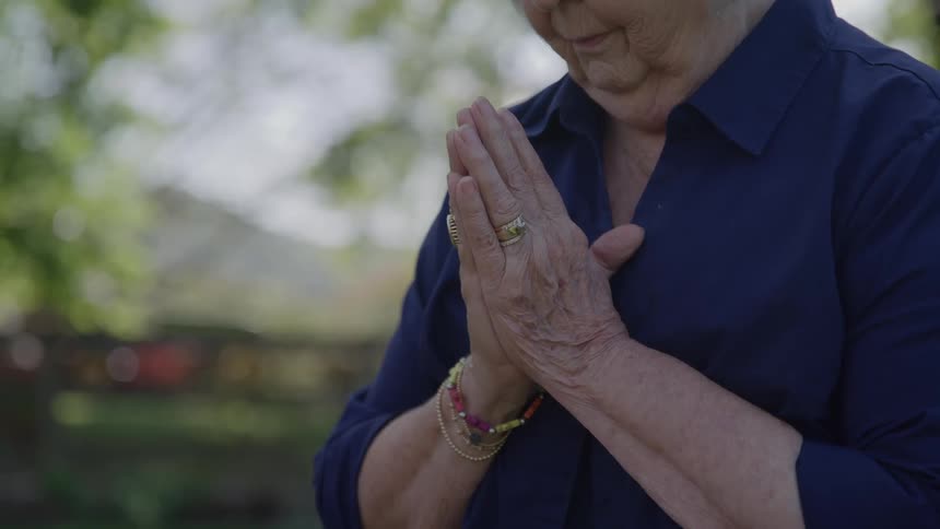 A elderly woman outdoors presses her hands together in prayer wearing a navy blouse and colorful bracelets looking peaceful and reflective