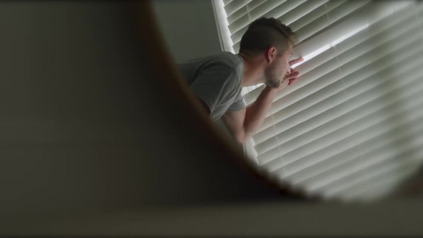 A man using his finger to open a piece of a venetian blind and look out the window