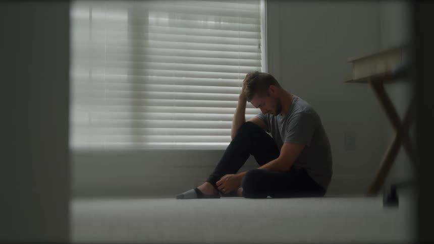 A young man sitting on the floor inside an empty room beside the window at midday