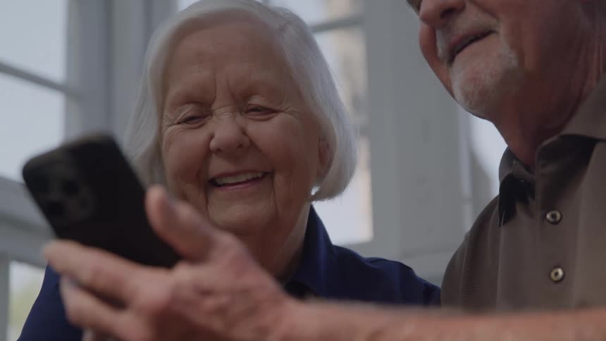 The face of an elderly woman smiling as she looks at a cell phone held by another person