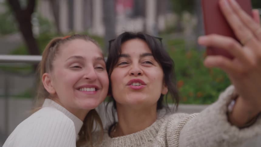 A two women smile brightly in cozy sweaters as they take a selfie with the woman on the left flashing a peace sign in an urban park setting