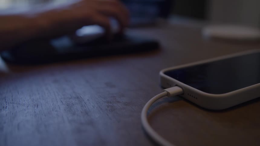 A smartphone in a white case charges on a wooden desk while a hand works at a computer in the blurred background