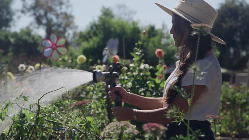 A woman wearing a hat is using a hose to water the plants in her yard