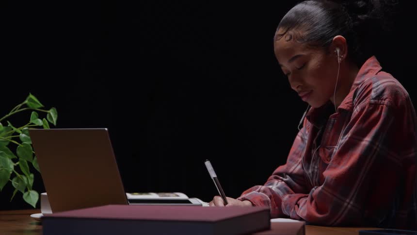 A young woman listens to earbuds while writing at a work desk