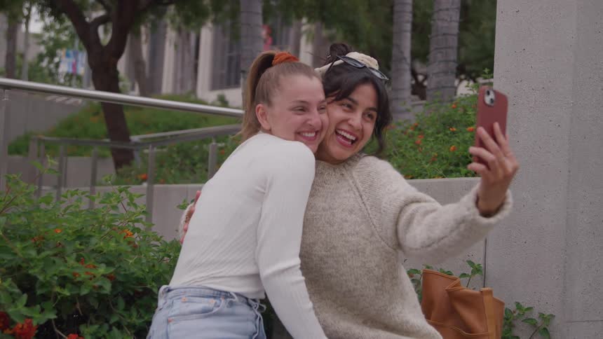 Two women smile while taking a selfie outdoors on a summer day