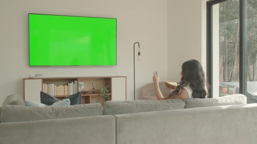 Woman walks into the living room carrying a bowl of popcorn and points the remote control at the television as she sits down on the couch