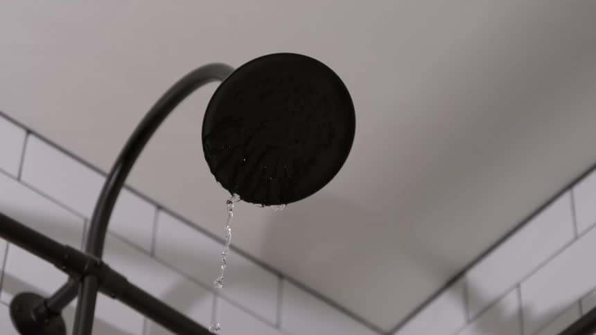 A lowangle closeup shows a matte black rain showerhead slowly dripping water in a modern bathroom the persistent drips fall against a backdrop of classic white subway tiles highlighting a small but continuous leak