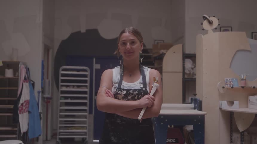 Woman with a paintstained apron stands with arms crossed in a pottery studio