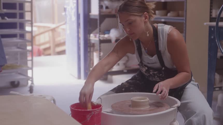 Woman shapes clay on a pottery wheel inside a brightly lit studio