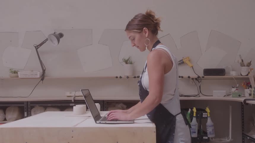 A woman looks focused and thoughtful as she types on a laptop in a pottery studio surrounded by shelves tools and clay supplies