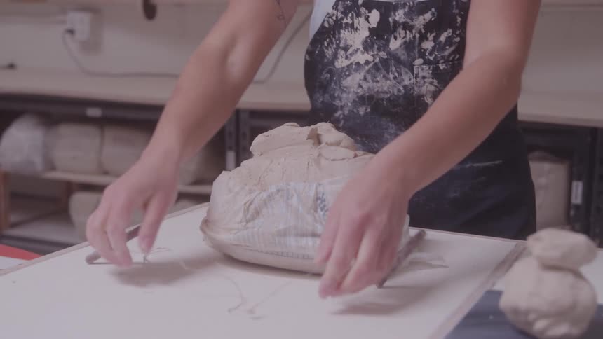A person in a splattered apron stands at a table in a studio using a tool to cut and shape a block of white clay