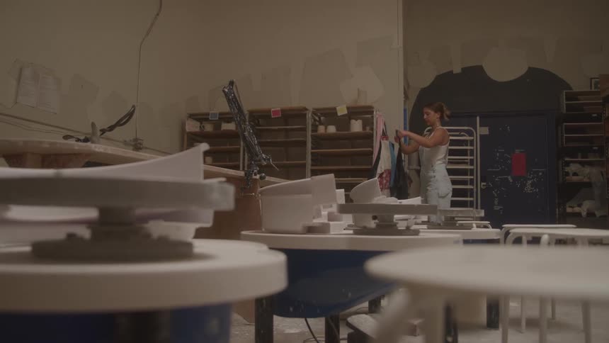 Young woman walks into a room with shelves and pottery wheels on tables and puts on an apron