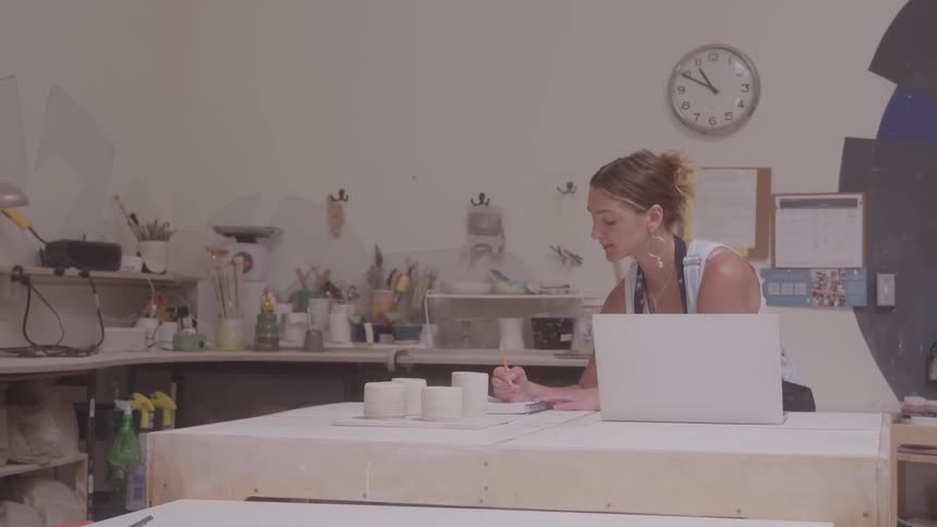 Young woman sits at a table with a laptop computer and ceramic cups as she writes on paper