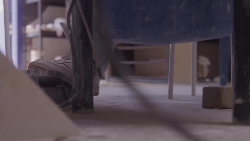 A lowangle shot on a dusty concrete floor focuses on the feet of a person in sneakers stepping on the lever of a pottery wheel the movement is quick and precise suggesting the beginning or end of the wheels rotation in a workshop setting