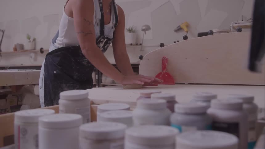 A medium shot shows a woman in a tank top and apron applying a reddish glaze to a piece of pottery in a bright workshop in the foreground numerous jars of glazes and paints are blurred framing the central action