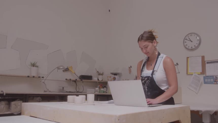 Young woman wearing a black apron walks into a room and types on a laptop computer
