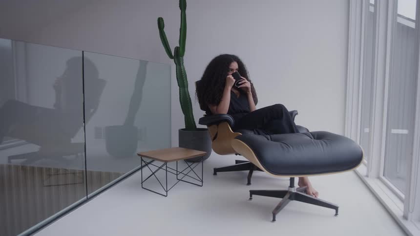 A young woman sitting on a chair on a balcony at midday drinking a cup of coffee