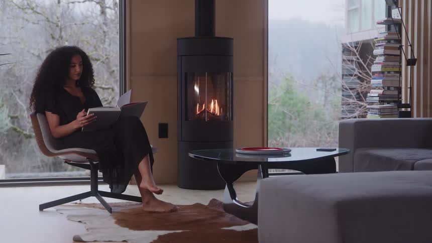 A woman with long dark curly hair is calmly sitting in a modern upholstered chair reading a book in a luxurious living room with a lit freestanding fireplace and expansive windows revealing a wooded exterior