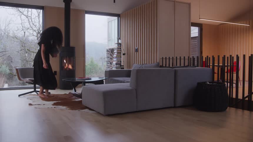 A woman walks barefoot carrying a folder through a modern cozy living room with wooden walls and large windows overlooking a misty landscape