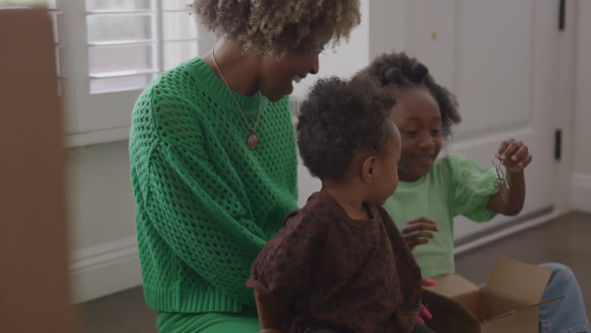 Mother smiles watching her daughter hold a set of keys as they all sit on the floor during the daytime