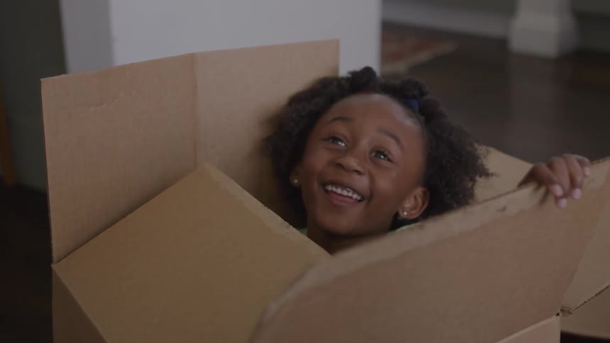 A black female child sits in a large box smiling and playing with the lid